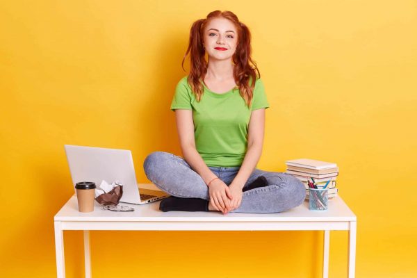 Young red haired female student sitting on white table with crossed legs, funny lady with ponytails studying, looking at camera, wearing casual clothing.